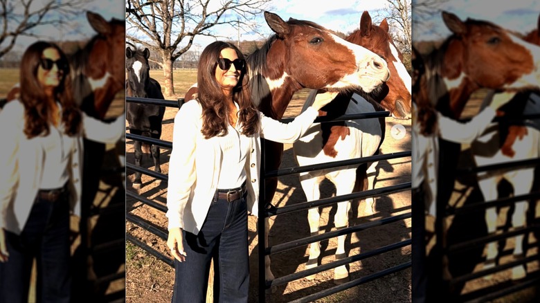 Kaitlan Collins poses next to horses wearing navy blue jeans and a white cardigan