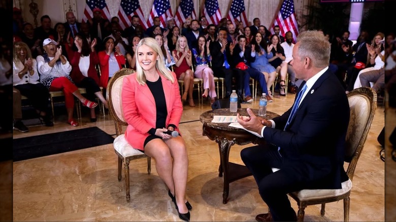 Karoline Leavitt sitting in a black minidress with a coral blazer