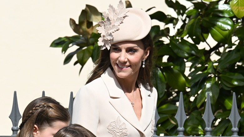 Kate Middleton, a middle-aged woman with pale skin and dark brown hair, is all smiles as she walks with her two children. She is wearing a cream blazer with floral appliques and a disc-shaped hat.