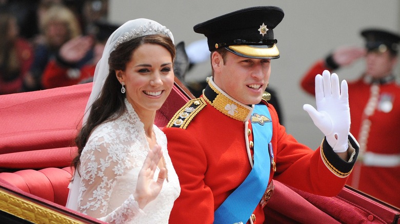Kate Middleton sits in a carriage beside Prince William. She is wearing a lacy white high-necked wedding gown with a tiara and veil. William is wearing a red captain's uniform and forage cap
