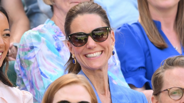 Kate Middleton wearing a blue top, sunglasses, and teardrop earrings smiling at Wimbledon 2026.