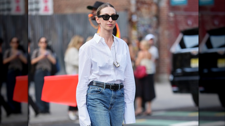 A woman in a white button down, jeans, and a silver necklace with a large pendant.