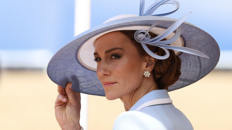 Kate Middleton looking to the side wearing an ornate blue hat and pearl flower-shaped earrings.