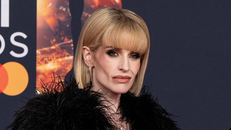 Kelly Osbourne at an event. She is a pale woman with defined cheekbones, heavy makeup, and a blonde bob haircut with blunt bangs. She is wearing a black feathered dress.