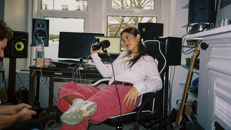Lola Consuelos wearing hot pink pants and a white top, sitting in a gaming chair holding a recording mic.