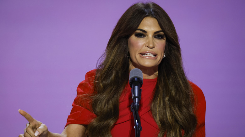 Kimberly Guilfoyle speaking at a podium with her long hair extensions