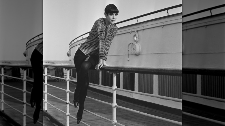 Peggy Moffitt posing on the railing of a ship wearing a checkered jacket with a black skirt and tights.