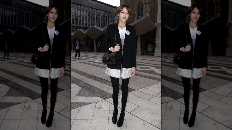 Alexa Chung photographed wearing a black jacket, white dress, black tights, and black shoes outdoors.