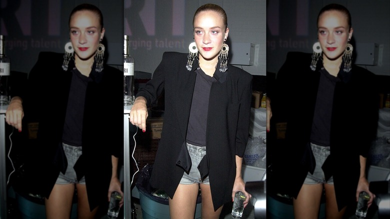 Chloë Sevigny photographed with denim shorts, black tee, oversized black blazer, and statement earrings with her hair slicked back.