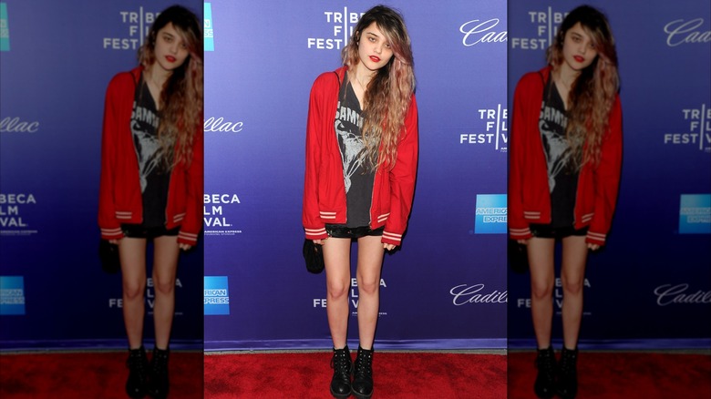 Sky Ferreira posing at a red carpet event with her head to the side wearing a red jacket, band shirt, black shorts, and combat boots.