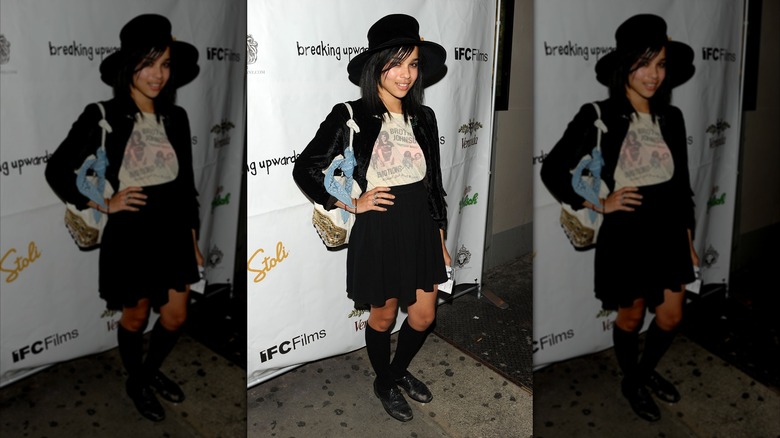 Zoë Kravitz posing at a movie premiere with her hand on her hips, wearing a black jacket and skirt with a white graphic tee and a black wide-brimmed hat.