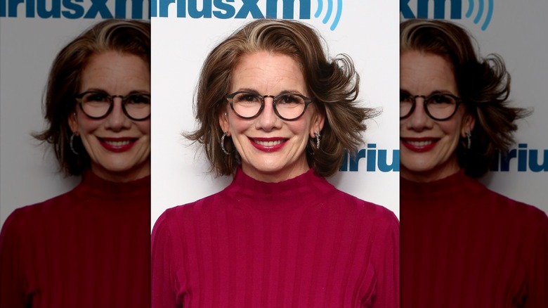 Melissa Gilbert smiles wearing a red turtleneck and round glasses