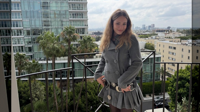 Skywalker Hughes smiles in a gray coat and skirt on a balcony.