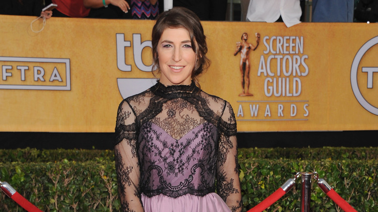 Mayim Bialik smiling on the red carpet wearing a lilac gown with a black-lace overlay top.