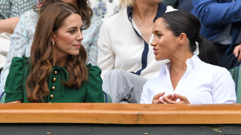 Meghan Markle in a white shirt sitting next to Princess Kate wearing a green shirt.