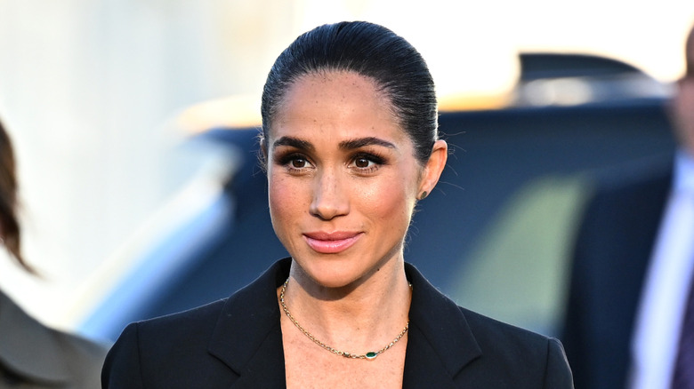 Meghan Markle smiles with her hair in a slicked-back bun wearing a black blazer
