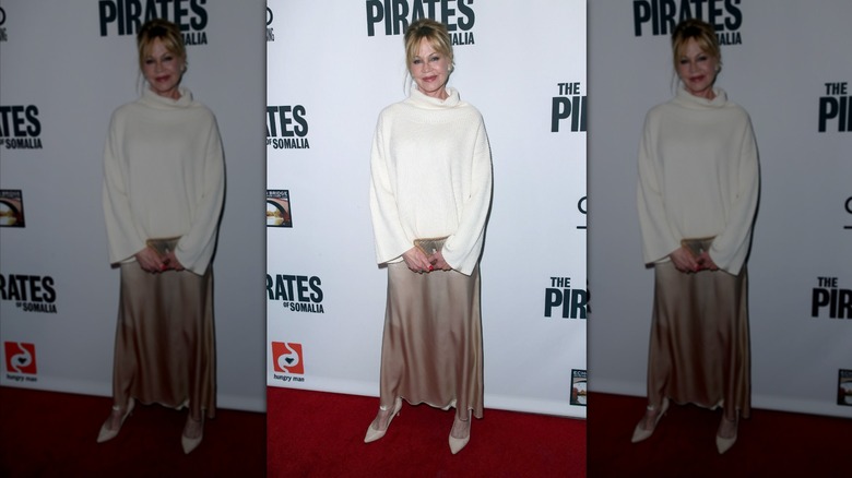 Melanie Griffith smiles wearing a white turtleneck and a beige satin skirt