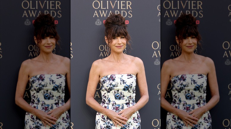 Minnie Driver posing at an event is a strapless dress with a floral pattern and her hair in an updo.