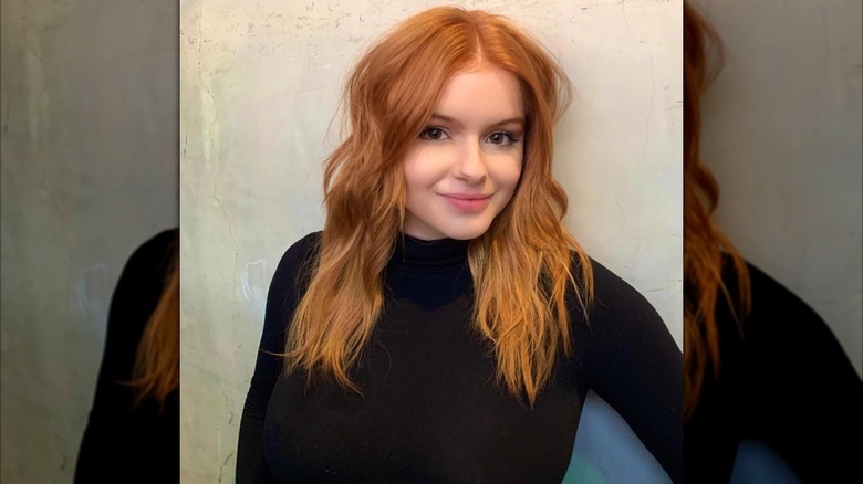 Ariel Winter posing in a black turtle neck with wavy red hair against a light backdrop.