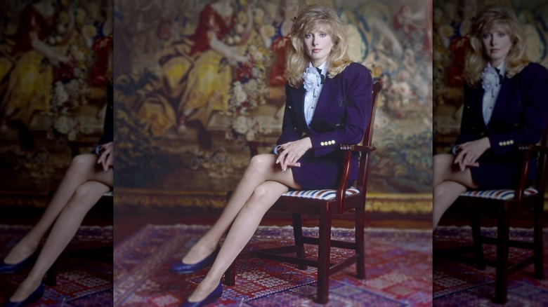 Morgan Fairchild sitting in a chair with crossed legs wearing a dark-purple skirt suit.