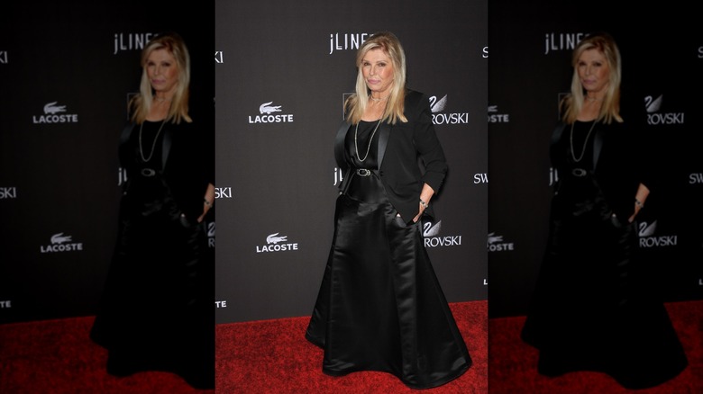 Nancy Sinatra posing at a red carpet event with her hands in her pockets wearing a black silky skirt and a black blazer.