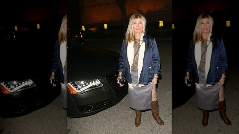 Nancy Sinatra posing at an event wearing a silver skirt and sparkly gold top with a dark blue coat and brown boots.