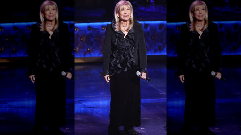 Nancy Sinatra holding a microphone onstage wearing a black ruffled top and a black skirt.