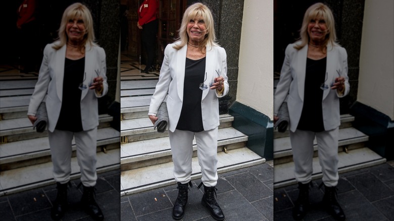 Nancy Sinatra photographed outside holding her glasses and wearing white pants an blazer with a black shirt and combat boots.