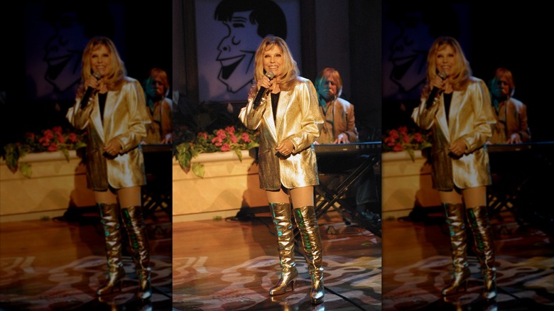 Nancy Sinatra performing on stage holding a microphone wearing gold knee-high boots and a gold blazer.