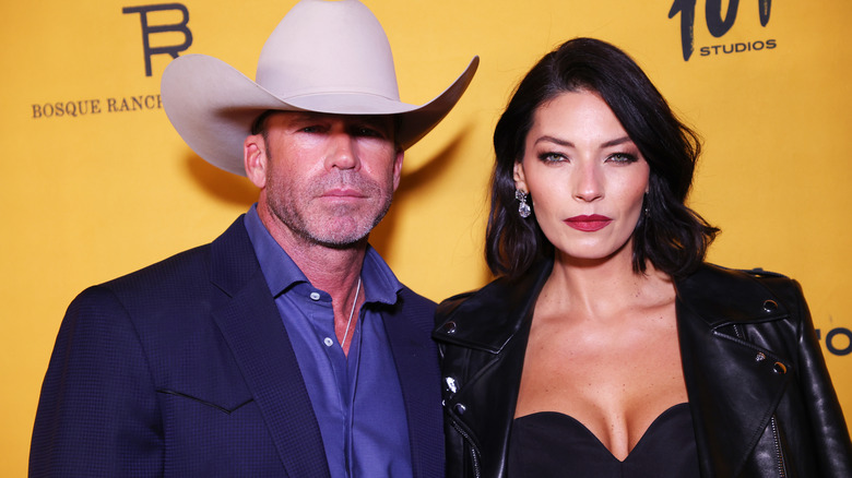 Taylor Sheridan and wife Nicole Sheridan pose on the red carpet together in front of a gold back drop.