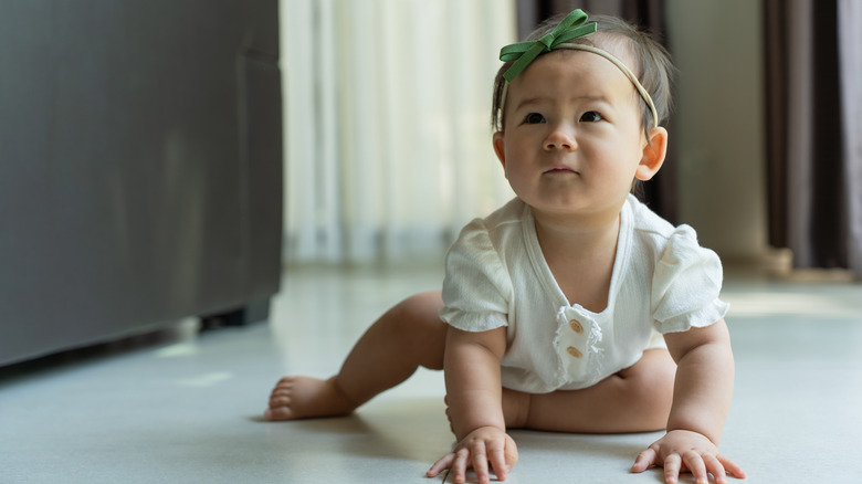 A baby crawling while wearing a headband and a white romper