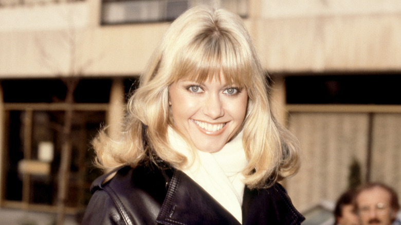 Olivia Newton-John smiles for photographers in a black leather jacket and white wool scarf