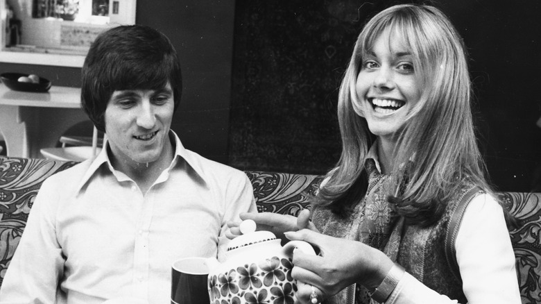 Bruce Welch and Olivia Newton-John are seen having tea together on a couch in a black and white photo