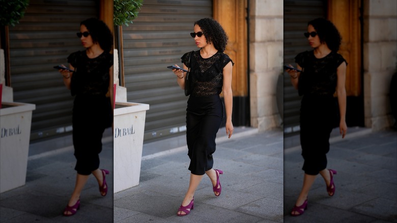 A woman walking down the street in a black pencil skirt, lacey black top, and purple block heels.