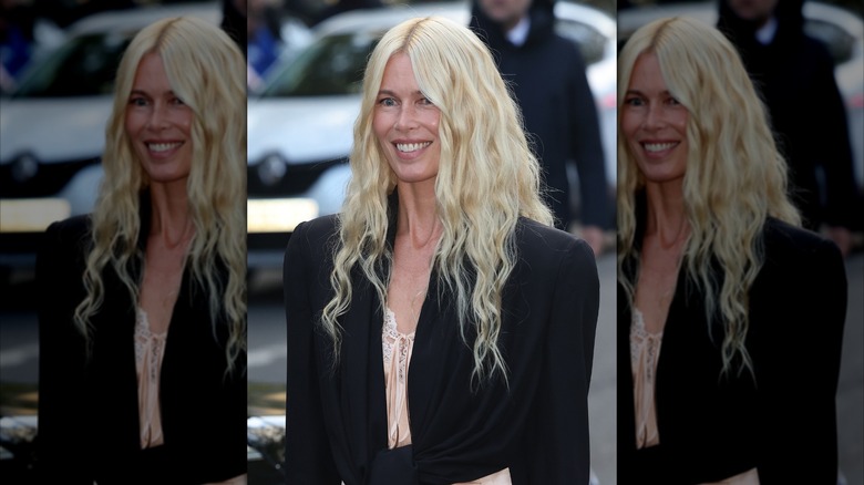 Claudia Schiffer smiling in a white top and black coat with blond hair in mermaid waves.