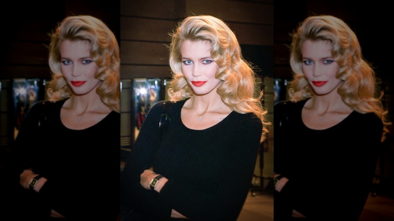 Claudia Schiffer posing in a black scoop-neck top with red lipstick with her arms crossed and curly blond hair worn down.