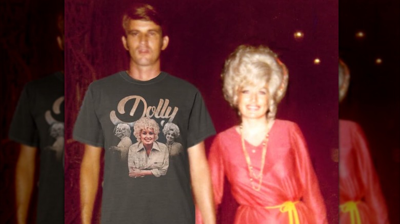 Dolly Parton smiles while holding hands with husband Carl Dean, who is photoshopped to be wearing one of her merch T-shirts.