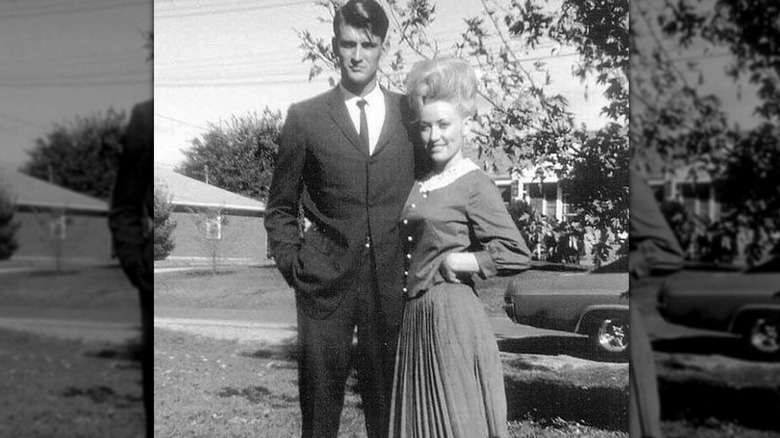 A black-and-white photo of Carl Dean and Dolly Parton posing together outside in front of an old fashioned car.