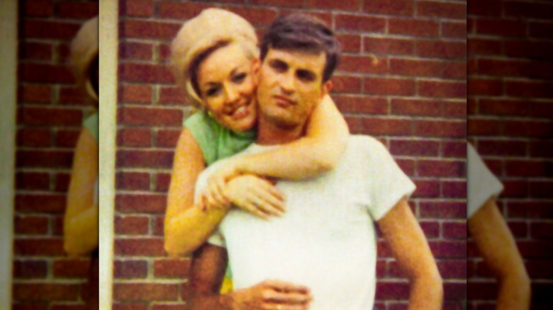 A young Dolly Parton is pictured in front of a brick wall while hugging husband Carl Dean from behind.
