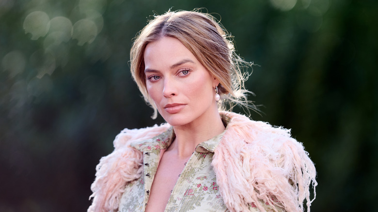 Margot Robbie posing outside at an event with her hair up wearing a detailed, fuzzy outfit.