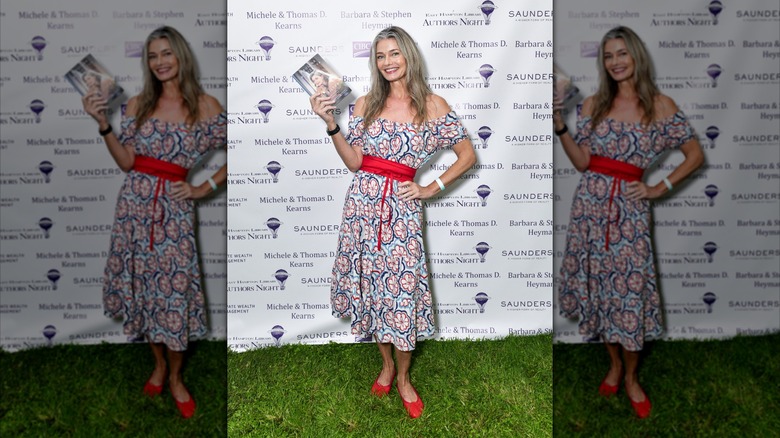 Paulina Porizkova standing on grass holding a book and wearing red shoes and a dress at an event.