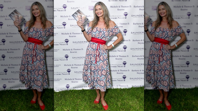 Paulina Porizkova standing on grass holding a book and wearing red shoes and a dress at an event.