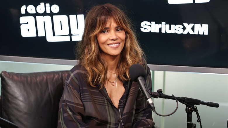 Halle Berry wearing a brown plaid shirt sitting in front of a microphone for a podcast taping.