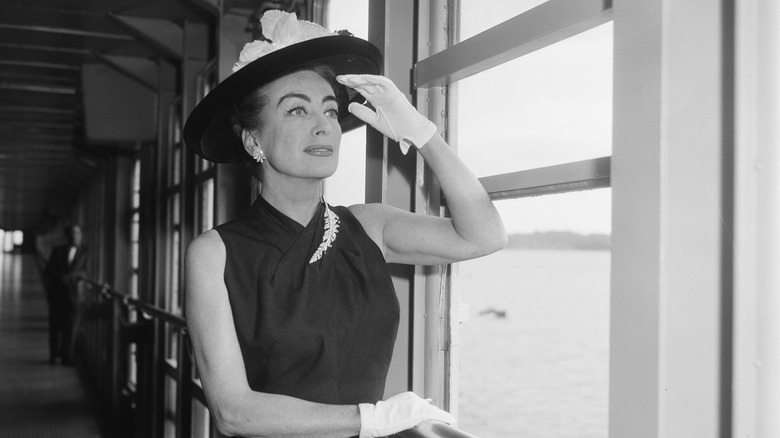 Joan Crawford looking out the window of a ship wearing a wide-brimmed hat, black dress, and white gloves.