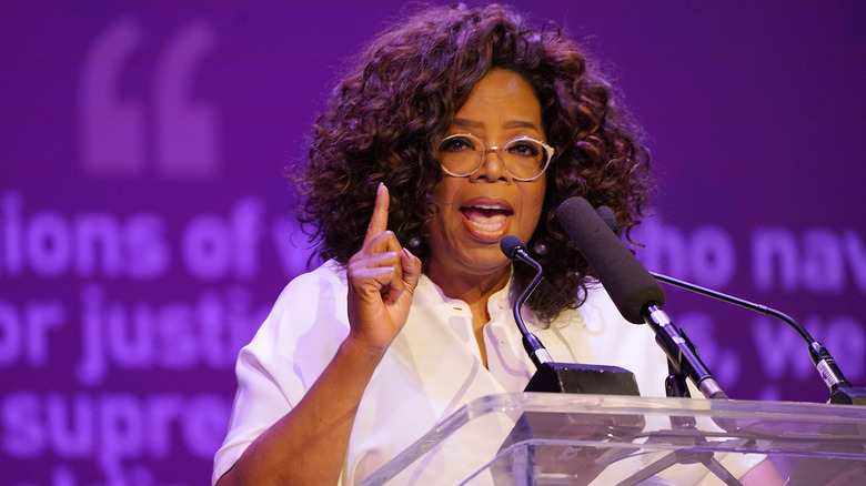 Oprah Winfrey speaks into a microphone on stage in front of a purple backdrop.