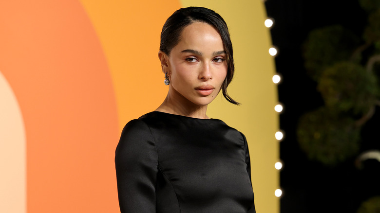 Zoë Kravitz poses in front of an orange and yellow backdrop while wearing a black dress and an updo.