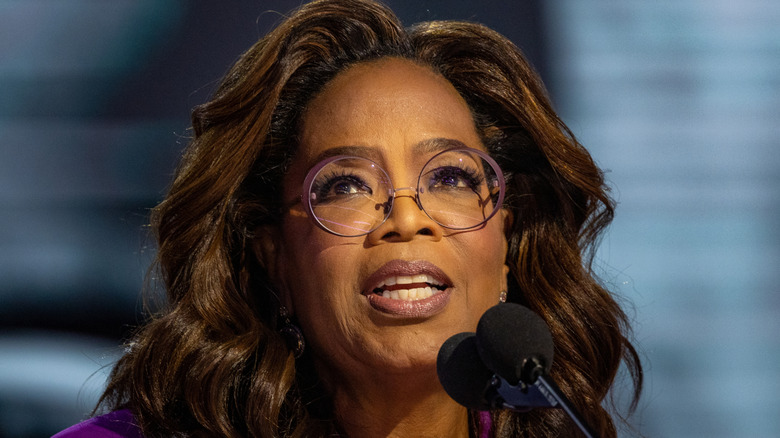 Oprah Winfrey speaks onstage in a purple blazer and purple round frame glasses.