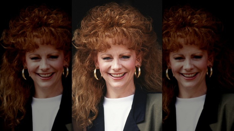 Reba McEntire with large, curly red hair wearing a white top and a blazer in 1990.