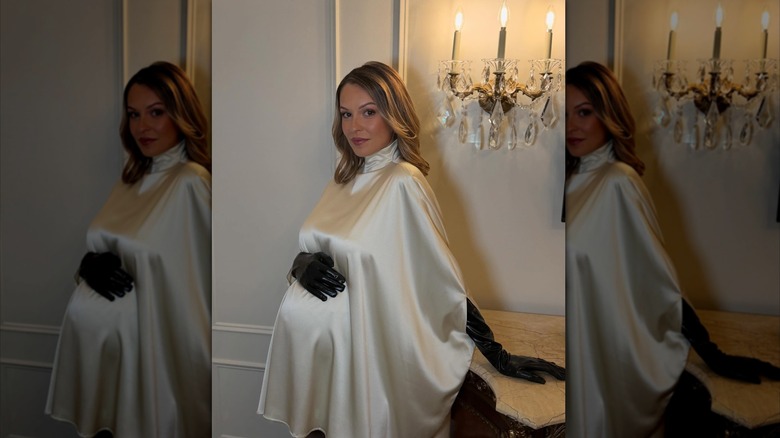 Kayla Wallace wearing a white silk turtleneck dress and black leather gloves with her hand on her baby bump.