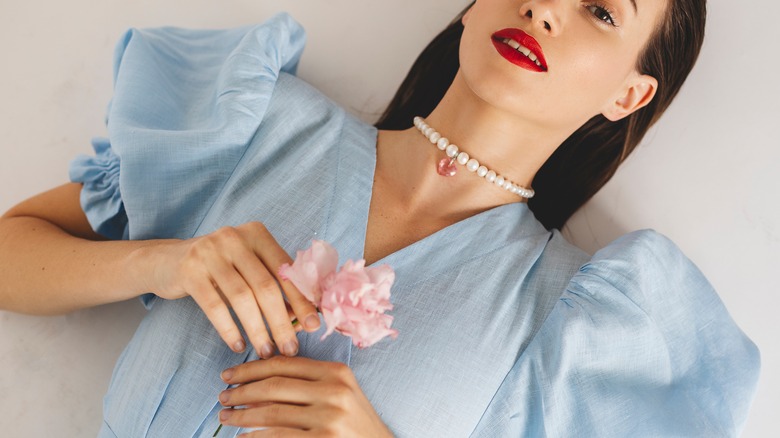 woman lays on the ground in blue puff sleeve dress and pearl necklace holding a pink rose